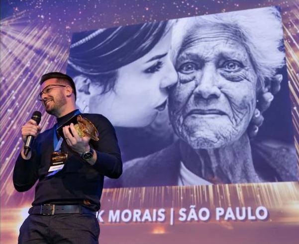 Fotógrafo recebendo prêmio no palco com foto premiada no telão